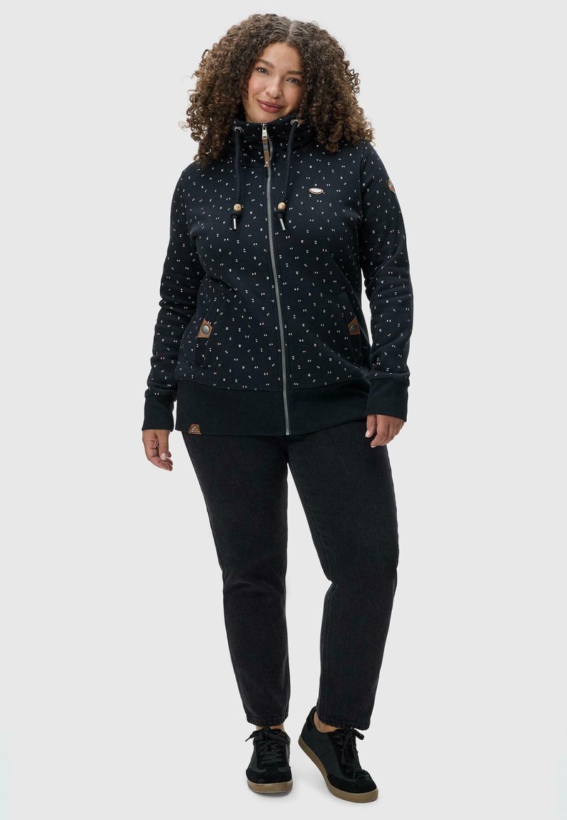 Sudadera negra con cremallera y lunares blancos, capucha con cordón y dos bolsillos laterales. Combinada con jeans oscuros y zapatillas negras.