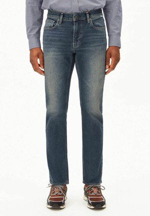 Jean en denim bleu foncé légèrement délavé, coupe droite, design à cinq poches, fermeture par bouton, associé à des chaussures de sport.