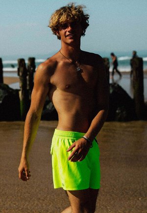 Jeune homme torse nu aux cheveux bouclés portant un short de bain vert néon, debout sur une plage ensoleillée avec des poteaux en bois et des vagues de l'océan en arrière-plan.