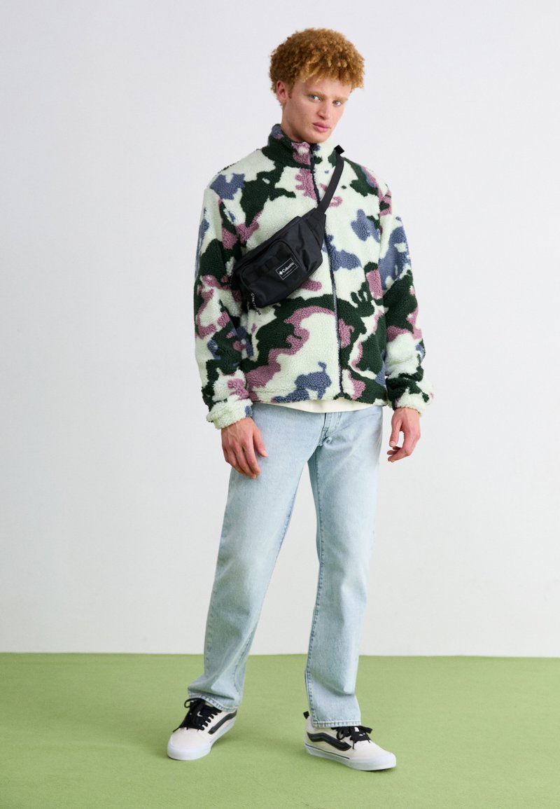 Casaco de fleece com padrão de camuflagem verde, rosa e azul, bolsa de cintura preta, jeans azul claro e sapatilhas preto e branco num chão verde.