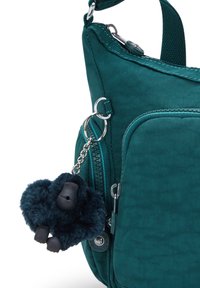 Borsa a spalla in tessuto teal con tasca frontale con zip e un portachiavi di gorilla in peluche nero attaccato a un anello metallico sul lato.