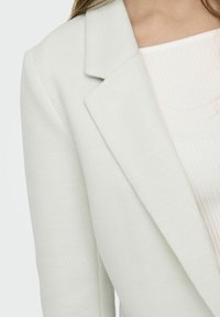 Lichtgrijze blazer met een subtiel visgraatpatroon, gestructureerde revers en een gladde textuur. Gelaagd over een geribbeld wit top.