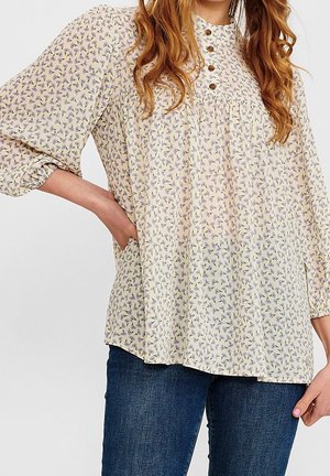 Frau trägt eine locker sitzende, langärmelige Bluse mit Blumenmuster und braunen Knöpfen, kombiniert mit dunkelblauen Jeans, Hand ruht auf der Hüfte.