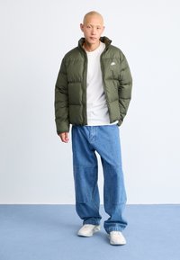 Puffer jacket in olive green with a zip front and high collar, worn over a white shirt, paired with blue loose-fit jeans and light shoes.