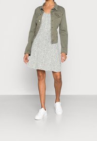 Mujer vestida con un vestido ligero con estampado, chaqueta verde oliva de botones y zapatillas blancas, de pie sobre un suelo blanco liso y con fondo blanco.