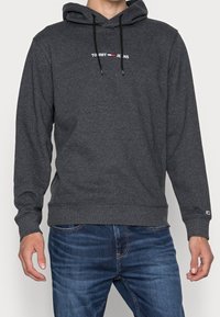Homme portant un hoodie Tommy Jeans gris foncé avec des cordons de serrage noirs et un jean en denim bleu, debout avec les bras relâchés sur les côtés.