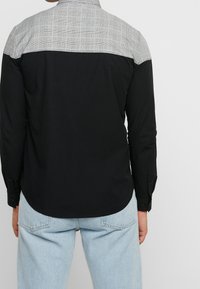 Långärmad skjorta med svart kropp och grå rutig axelparti, med knäppta manschetter och en rundad nederkant. Slät textur hela vägen igenom.