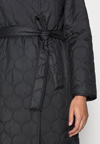 Gepolsterter schwarzer Mantel mit kreisförmigem Nähmuster, Stoffgürtel, der in der Taille gebunden ist, und die rechte Hand entspannt an der Seite.