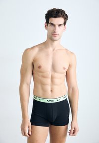 Schwarze Nike Boxershorts mit einem hellgrünen Bund, auf dem das Logo abgebildet ist. Der Stoff hat eine glatte Textur und ein enges Design.