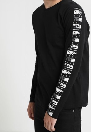 Person iført en sort langærmet skjorte med hvidt grafisk tekst "STAY BOLD" og abstrakte mønstre, der løber ned ad højre ærme.