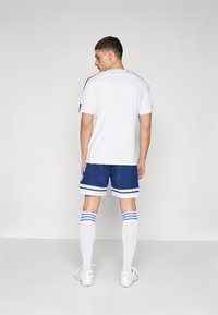 Witte sportshirt met zwarte accenten, gecombineerd met marineblauwe shorts met witte en blauwe strepen, witte kniekousen en sportschoenen.