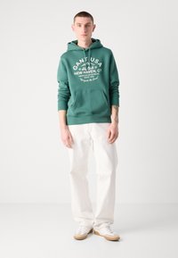 Hanorac verde cu buzunar tip marsupiu, text alb cu logo și manșete cu nervuri. Asortat cu pantaloni albi largi și adidași albi.