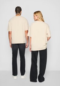 Beige oversized tričká s krátkym rukávom a voľným strihom nosené s čiernymi nohavicami, zobrazené na jednoduchom pozadí.