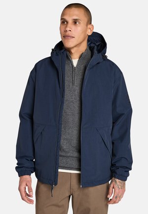 Mand i en marineblå hættetrøje over en grå sweater og kaki bukser, med tatoveringer synlige på hans hånd. Neutral baggrund.