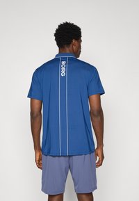 Muška plava polo majica s bijelim prugama, logo "BORG" na leđima, kratki rukavi i ovratnik, u kombinaciji sa sivim šortsevima.