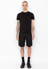 Zwarte crew-neck t-shirt, zwarte cargoshorts met zijzakken, en enkelsokken. Model draagt zwarte veterschoenen. Tatoeages zichtbaar op de armen.