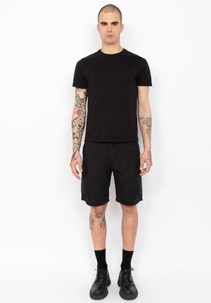 Zwarte crew-neck t-shirt, zwarte cargoshorts met zijzakken, en enkelsokken. Model draagt zwarte veterschoenen. Tatoeages zichtbaar op de armen.