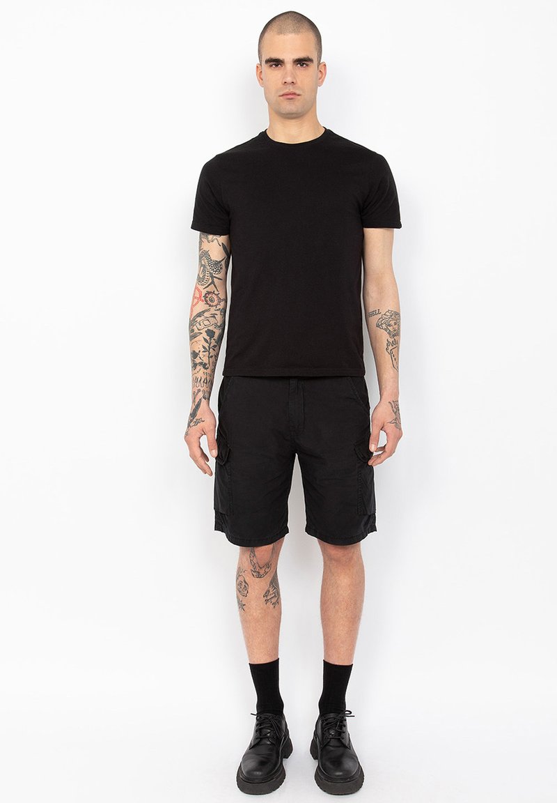 Zwarte crew-neck t-shirt, zwarte cargoshorts met zijzakken, en enkelsokken. Model draagt zwarte veterschoenen. Tatoeages zichtbaar op de armen.