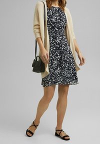 Kvinna i svart och vitt blommigt klänning, beige kofta, svart axelväska och svarta sandaler med remmar, stående framför en neutral bakgrund.