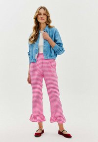 Modrá zkrácená džínsová bunda s květinovou výšivkou, bílý top, růžové kostkované kalhoty s volánky a červené ploché boty. Modelka stojí s rukama na bocích.