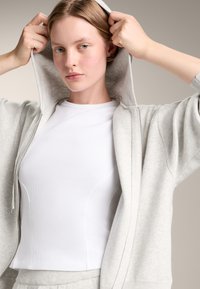 Svijetlo siva zip-up dukserica sa mekim teksturom, nosi se preko uske bijele rebraste majice. Dukserica ima kapuljaču i opuštene rukave.