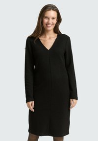 Schwarzes V-Ausschnitt-Kleid mit langen Ärmeln, strukturiertem Stoff, knielangem Schnitt und einer mittigen Nahtdetails, getragen über schwarzen Strumpfhosen.