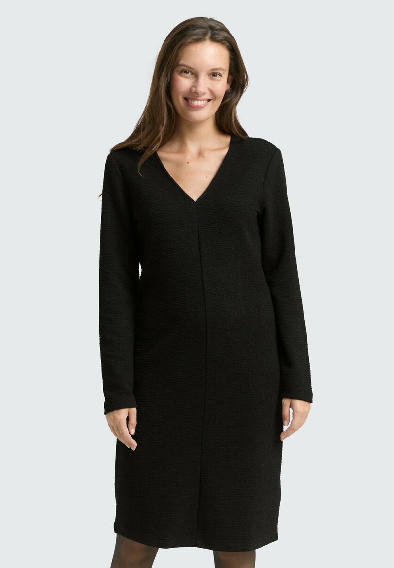 Schwarzes V-Ausschnitt-Kleid mit langen Ärmeln, strukturiertem Stoff, knielangem Schnitt und einer mittigen Nahtdetails, getragen über schwarzen Strumpfhosen.