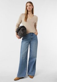Beige Langarm-Oberteil mit V-Ausschnitt, kombiniert mit weit geschnittenen hellblauen Jeans. Das Model hält eine schwarze geflochtene Handtasche. Braune Wildlederschuhe.