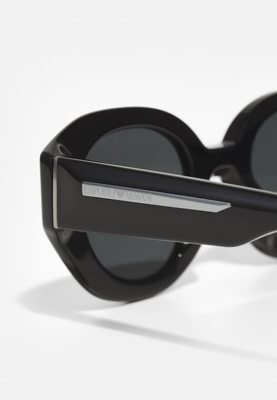 Ochelari de soare ovali negri cu lentile închise, având o ramă groasă din plastic și un accent metalic pe tâmple, gravat cu "Emporio Armani."
