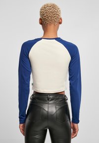 Gekort mouwloze shirt met blauwe raglanmouwen en een crèmekleurig lichaam. Beschikt over een ronde halslijn en zachte katoenen stof.