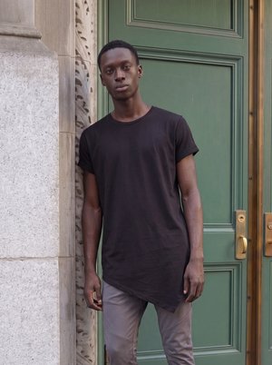 Junger Mann mit kurzen Haaren, der ein schwarzes asymmetrisches T-Shirt und graue Hose trägt, steht vor einer Steinmauer und einer grünen Tür.