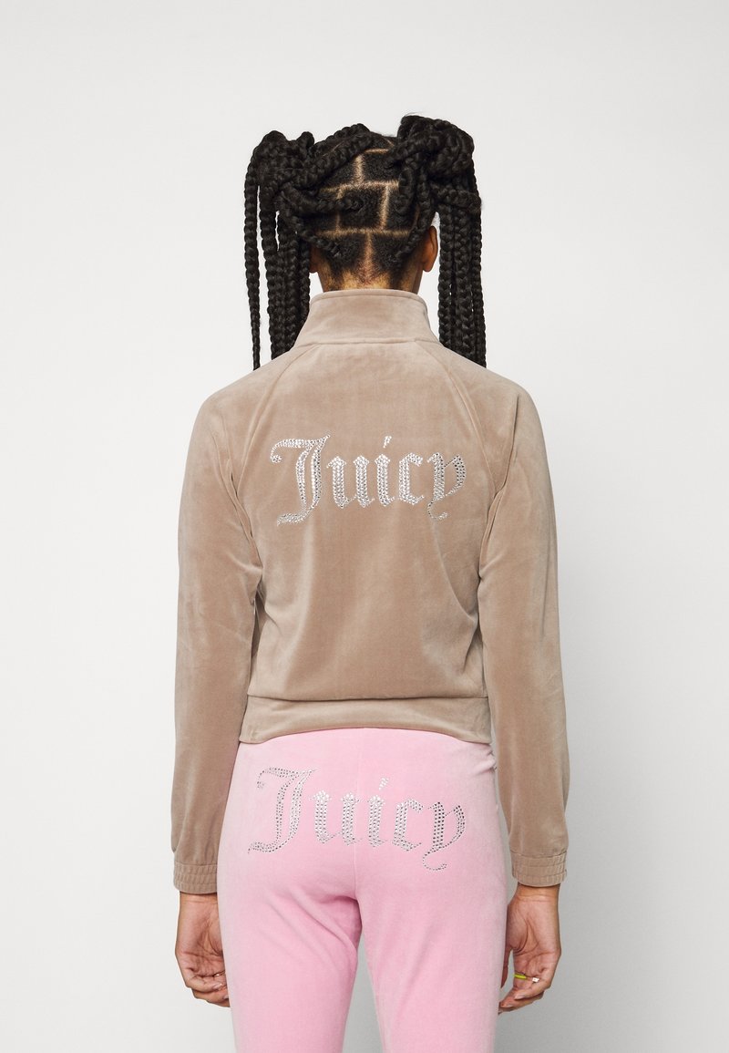 Velourträningsoverall i ljusbrunt med "Juicy"-design i rhinestones på baksidan av jackan och byxorna. Slät textur och tight passform.