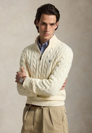 Jeune homme aux cheveux foncés portant un pull crème à torsades par-dessus une chemise rayée et un pantalon beige, debout les bras croisés.