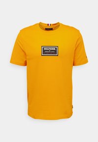 Světle žluté tričko s krátkým rukávem a černým obdélníkovým logem na hrudi, které zní "Hilfiger American Classic Est. 1985 New York".