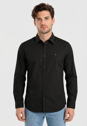 Jonge man met kort bruin haar, gekleed in een zwart overhemd met knopen en lichtblauwe spijkerbroek, staand tegen een effen lichtgrijze achtergrond.