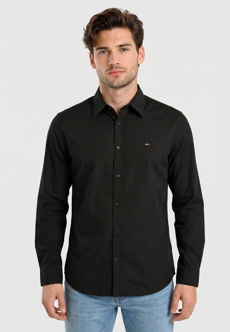 Giovane uomo con capelli corti castani che indossa una camicia nera con bottoni e jeans azzurri chiari, in piedi davanti a uno sfondo semplice grigio chiaro.