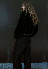 Veste en fausse fourrure noire à coupe décontractée, associée à un pantalon en denim foncé ample. De longs cheveux ondulés tombent sur une épaule.