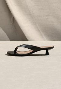 Sandale noire en cuir à talon en forme de tige avec un petit talon incurvé sur une surface beige neutre avec un fond en tissu.