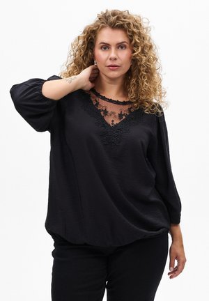 Zwarte blouse met volumineuze mouwen, kantdetails langs de halslijn, zachte textuur en een ontspannen pasvorm. Draagt met zwarte broek.