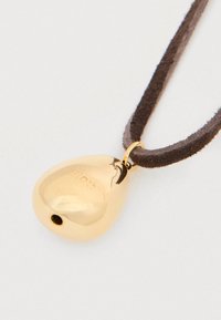 Χρυσό μενταγιόν σε σχήμα δακρύου σε καφέ κορδόνι από σουέτ. Λείο, αντανακλαστικό στέφανο με χαραγμένη λεπτομέρεια λογότυπου.