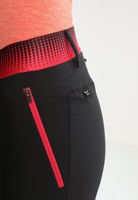 Zwarte sportbroek met een gestructureerde rode tailleband. Heeft een zijzak met een rode rits en een klein logo dicht bij de tailleband.