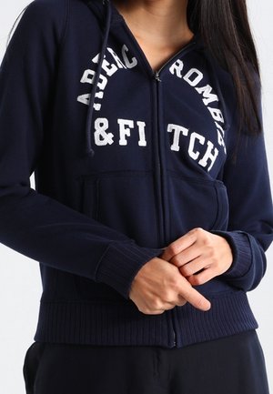 Navy zip-up hoodie with "Abercrombie & Fitch" text, front pockets, and drawstring hood worn by a person with hands clasped below chest.