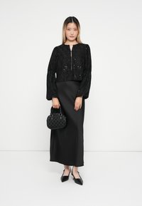 Črna vzorčasta kratka jakna s napihnjenimi rokavi, v kombinaciji z elegantno črno satensko maxi krilo. Drži teksturirano črno torbico in nosi nasvete čevlje.