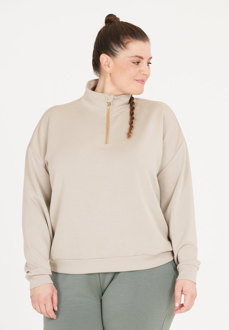 Beige sweatshirt med mock neck och halv dragkedja. Mjukt tyg med långa ärmar och avslappnad passform, kombinerad med gröna byxor.