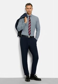 Hellblaues Hemd mit Kragen, kastanienroter, gestreifter Krawatte, dunkle Hose, schwarzer Gürtel und Schuhe; Jacke über eine Schulter gelegt.