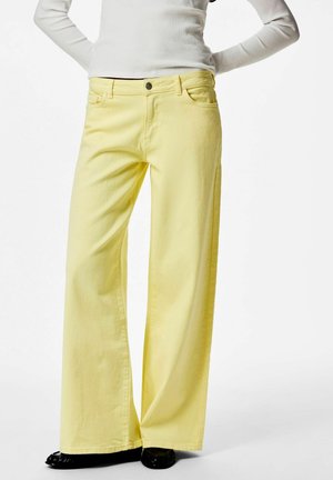 Persona che indossa pantaloni gialli a vita alta e gamba larga abbinati a un top bianco a maniche lunghe a coste e scarpe nere, in piedi con le mani dietro la schiena.