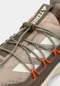 Chaussures de trail légères en beige et marron, avec dessus en mesh, accents réfléchissants, lacets élastiques et semelle texturée avec clips orange vif.