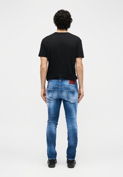 Άνδρας με σγουρά μαλλιά στέκεται γυρισμένος από πίσω, φορώντας απλό μαύρο μπλουζάκι, στενά μπλε τζιν με δερμάτινο ετικέτα και μαύρα παπούτσια.