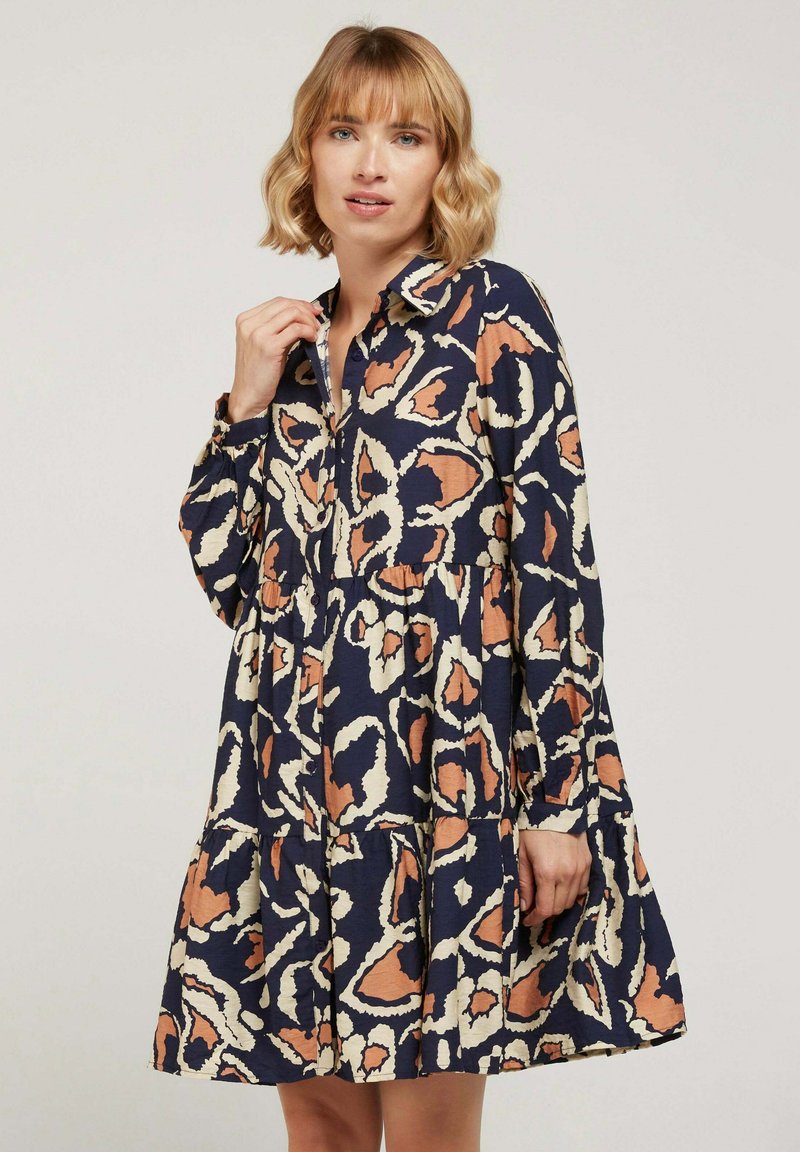 Robe de marine avec un design abstrait en beige et rouille, col boutonné, manches longues et jupe à volants. Le tissu semble texturé.