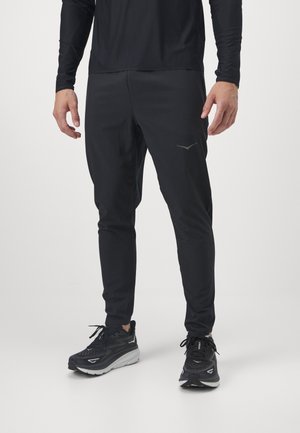 Mann trägt schwarze Sporthose, schwarzes langärmliges Oberteil und schwarze Laufschuhe und steht vor hellem Hintergrund.